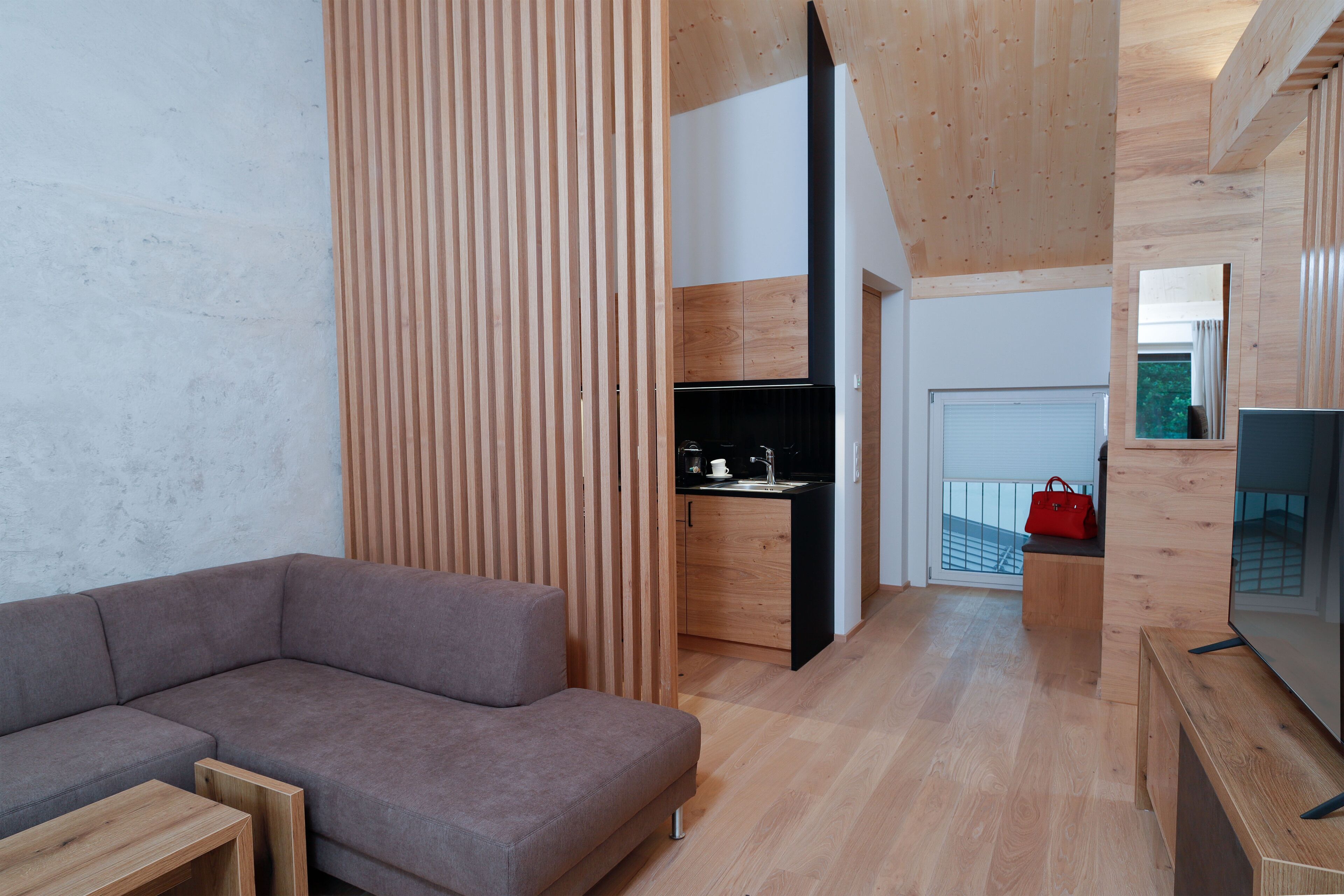 Un saló modern amb un sofà modular, una separació de fusta, una zona de cuina amb taulell negre i un televisor sobre un suport de fusta.