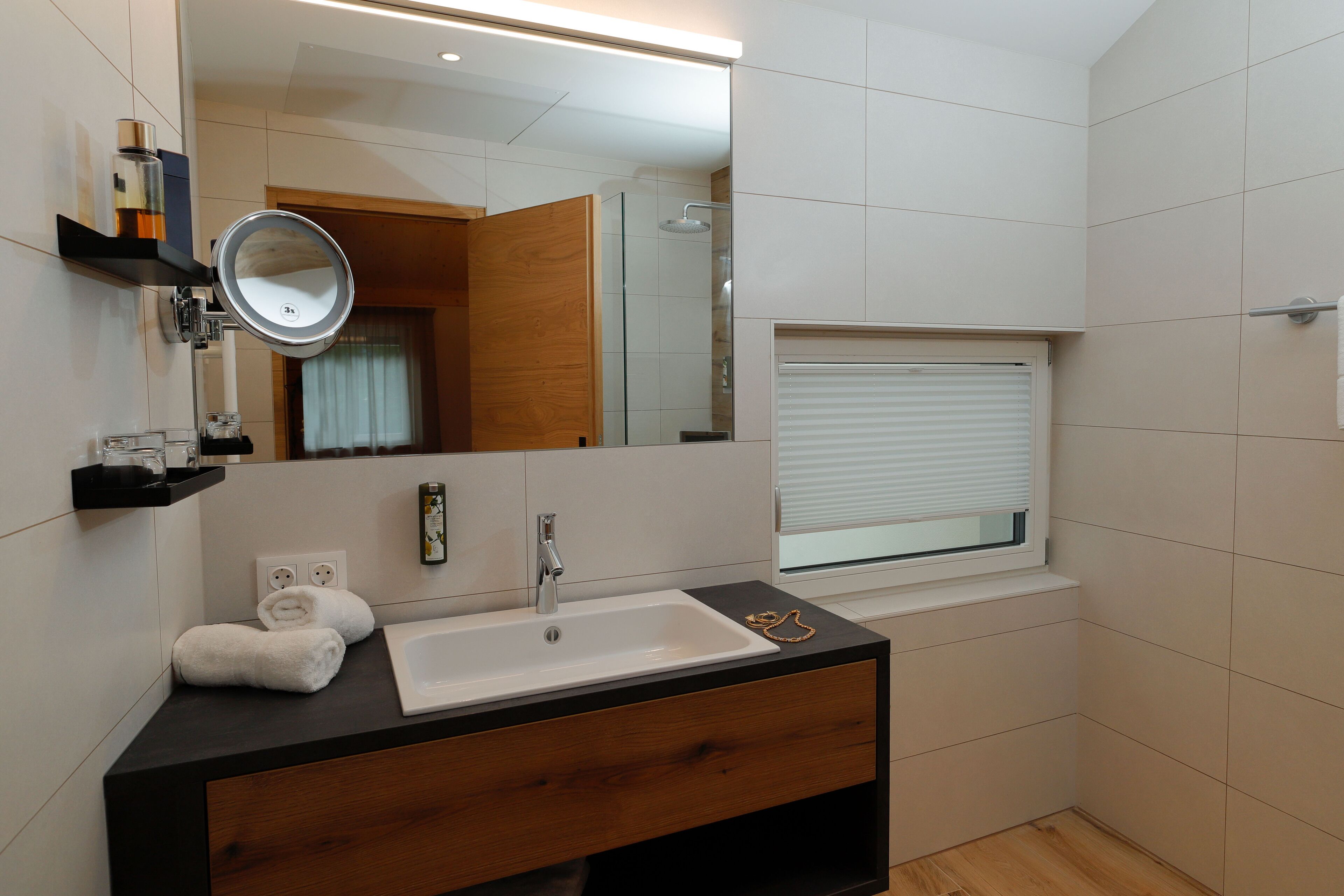 Un baño moderno con tocador de madera, un lavamanos blanco, un espejo y una ventana con persianas.