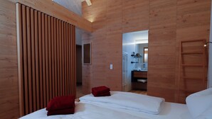 Quarto de hotel moderno com painéis de madeira, duas camas com roupa de cama vermelha e branca, um guarda-roupa de madeira e um espelho refletindo um banheiro com chuveiro e pia.
