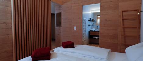 A modern hotel room with wooden paneling, two beds with red and white linens, a wooden wardrobe, and a mirror reflecting a bathroom with a shower and a sink.