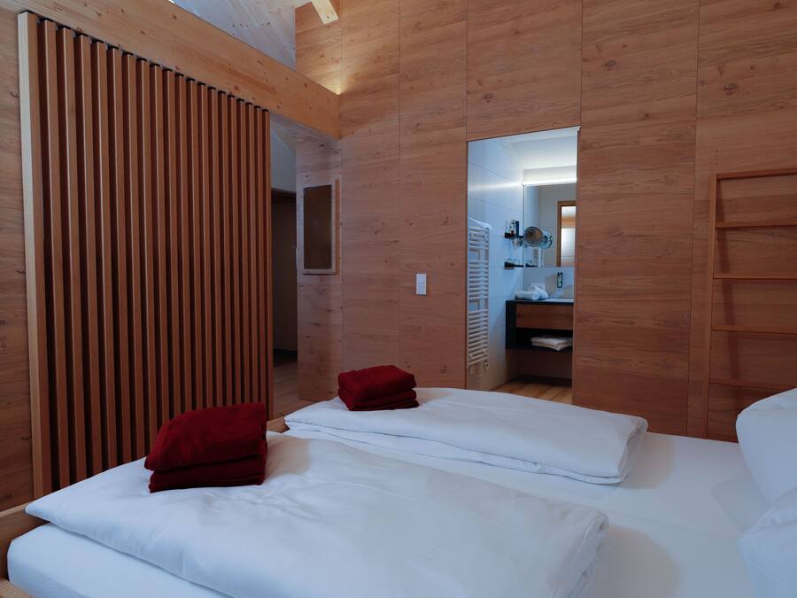 Ein modernes Hotelzimmer mit Holzvertäfelung, zwei Betten mit rot-weißer Bettwäsche, einem Holzschrank und einem Spiegel, der ein Badezimmer mit Dusche und Waschbecken reflektiert.