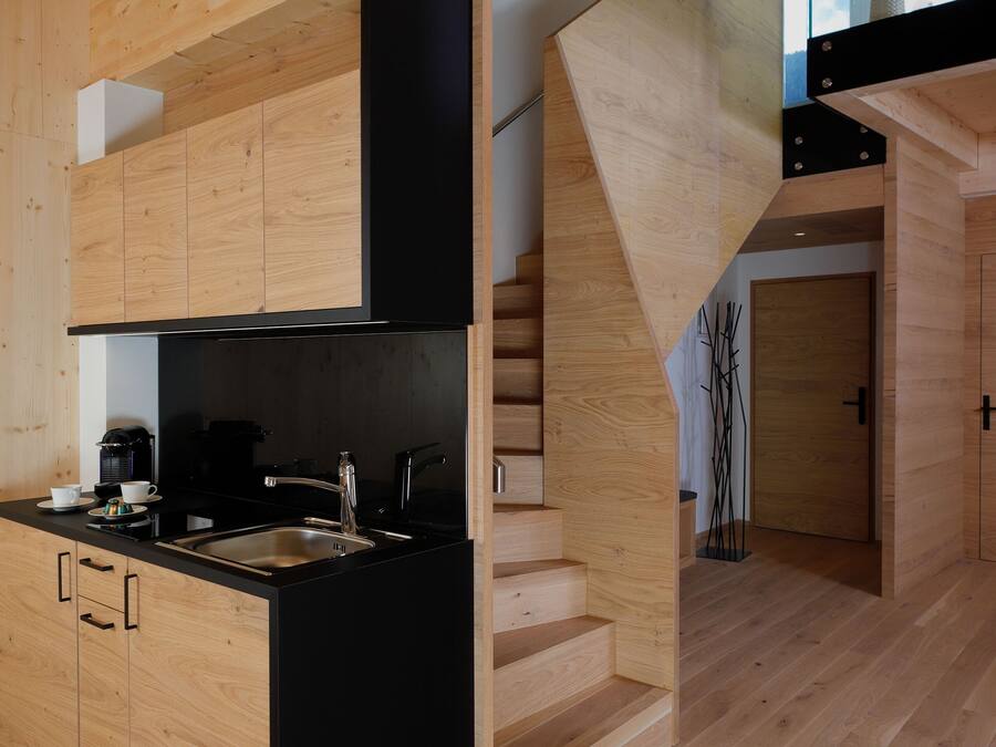 A modern kitchen with wooden cabinetry and a black countertop, a staircase with a glass railing, and a view of the outdoors.