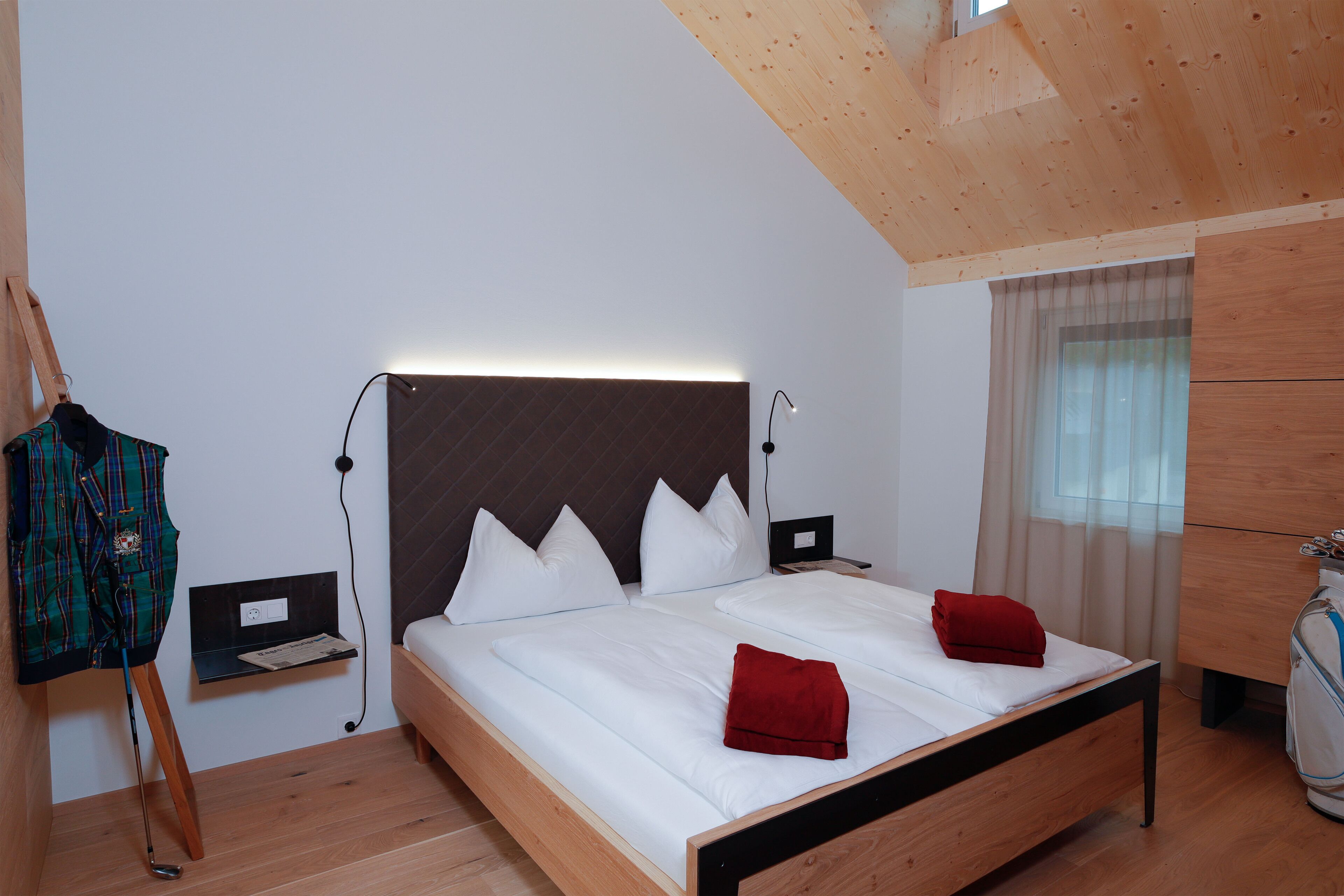 A modern bedroom with a wooden ceiling, a bed with white bedding and red pillows, a nightstand with a lamp, a coat rack, and a window with curtains.