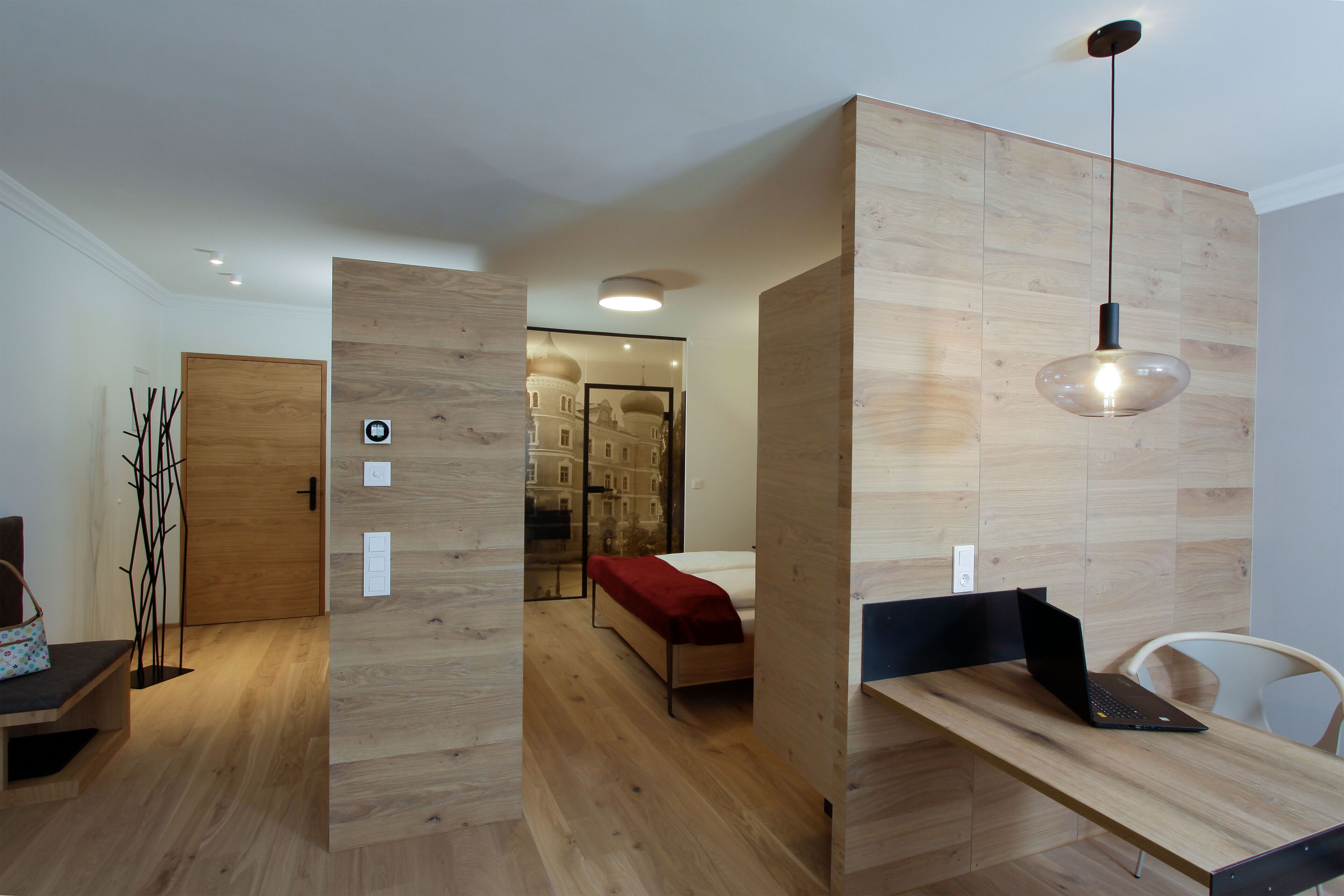 Una habitación moderna con suelo de madera, una cama con una colcha roja, un escritorio con un ordenador portátil y una lámpara colgante.