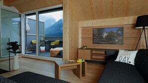 Uma sala de estar moderna com um sofá, uma mesa de centro e uma TV fixada na parede. Há uma janela com vista para montanhas e uma porta que dá para outro quarto.