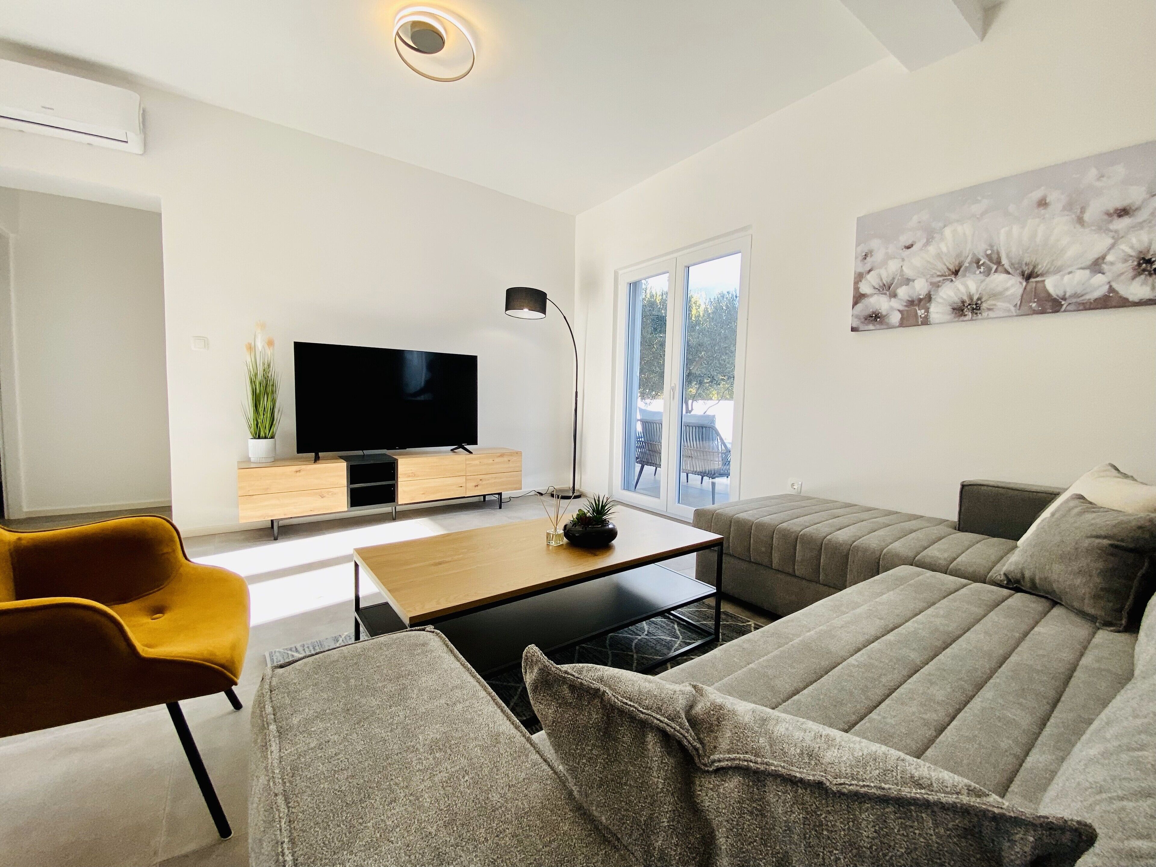 A modern living room with a flat-screen TV, a sofa, a yellow chair, and a dining table.