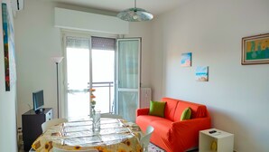 Een woonkamer met een rode bank, een eettafel met stoelen, een witte radiator en een raam met zonwering.