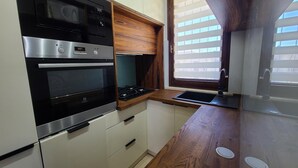 Cocina compacta con horno, microondas y estufa integrados. Cuenta con muebles de madera y una ventana con persianas.