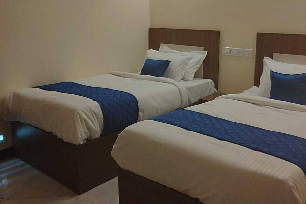 A hotel room with two single beds, each with a wooden headboard, white bedding, and blue accents. There is a wall-mounted light fixture above the beds and a framed picture on the wall.