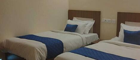 A hotel room with two single beds, each with a wooden headboard, white bedding, and blue accents. There is a wall-mounted light fixture above the beds and a framed picture on the wall.