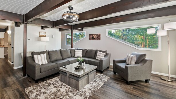 Un salon moderne avec un canapé d’angle, un petit fauteuil, une table basse et une grande fenêtre donnant sur un paysage verdoyant.