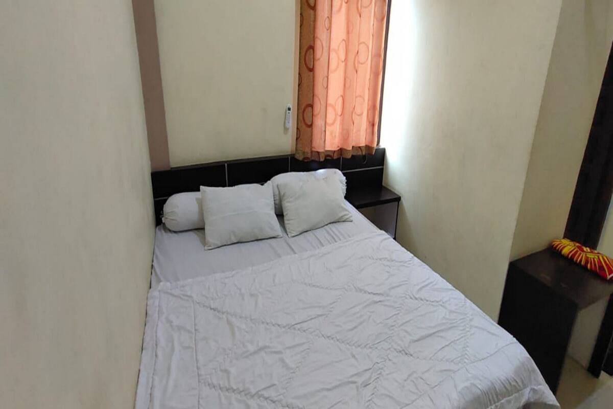 Ein Einzelbett mit weiß gestickter Bettdecke und zwei Kissen, ein Nachttisch mit Lampe sowie ein Fenster mit orangenen Vorhängen.