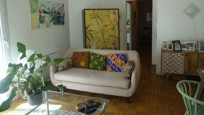 Een woonkamer met een bank, een salontafel en een eethoek met een stoel. Er zijn planten, een schilderij en een kast met foto's.