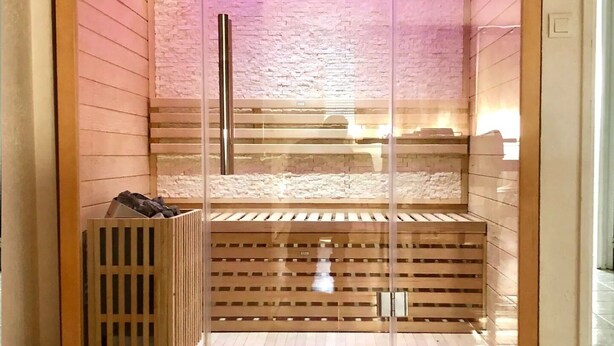 Una sauna con paredes de vidrio, interior de madera e iluminación rosada.