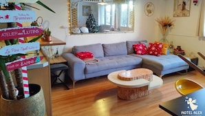 Una sala de estar acogedora con un sofá gris, una mesa de centro redonda y un espejo decorativo. Hay decoraciones navideñas, incluyendo un árbol pequeño y guirnaldas. Se ve un letrero con mensajes festivos.