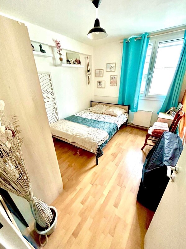 Un dormitorio compacto con cama, ventilador de techo, ventana con cortinas y una pequeña mesa con silla.