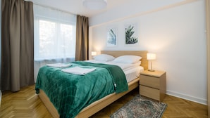 Een net opgemaakt bed met een groene deken, een nachtkastje met een lamp, en twee aan de muur hangende foto's.