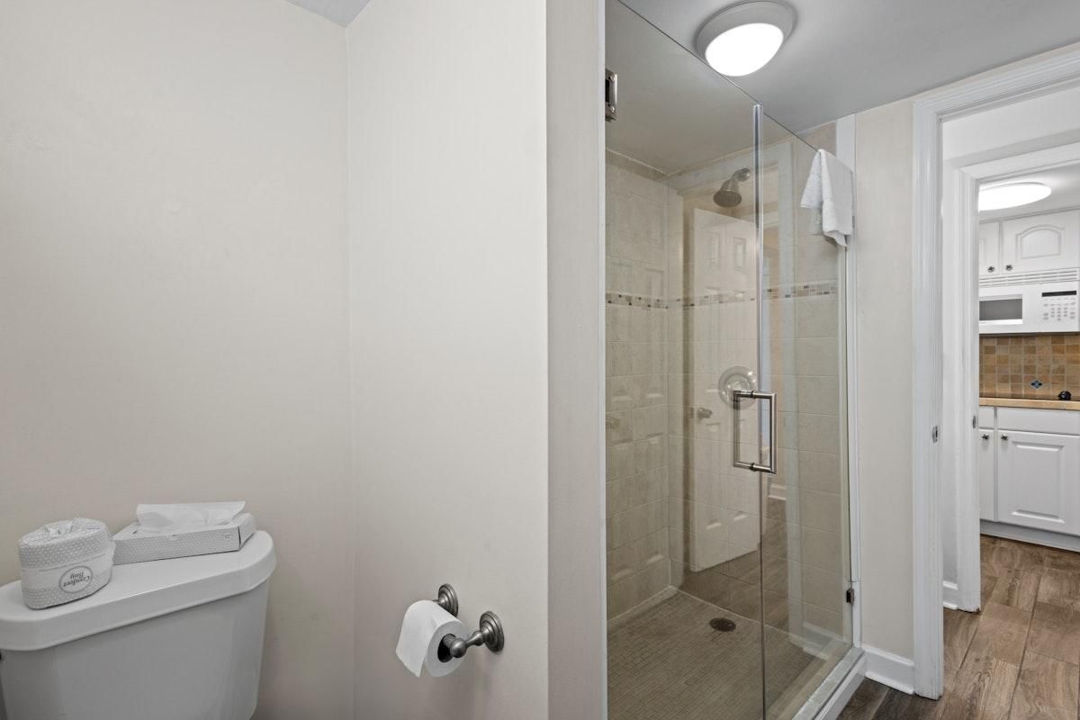 A bathroom with a toilet, a glass shower enclosure, and a visible kitchen area with cabinets.