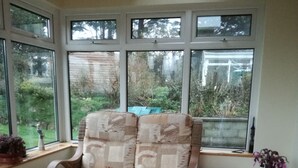 Une pièce baignée de lumière, avec une grande fenêtre, un fauteuil en osier, une petite table et un tapis à motifs.