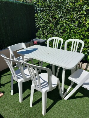Une table et des chaises en plastique blanc sont disposées sur une pelouse artificielle, avec une haie en arrière-plan.