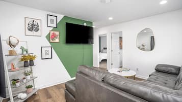 A modern living room with a grey leather sofa, a flat-screen TV mounted on a green wall, and a round mirror on the right.