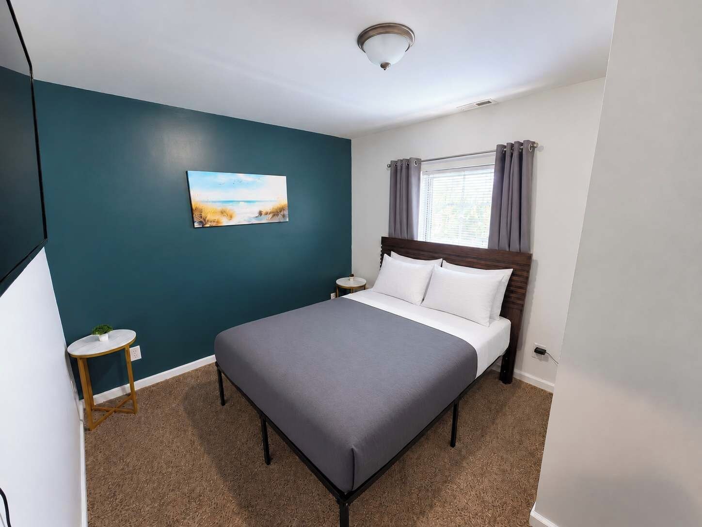 A hotel room with a bed, a small bedside table, a nightstand, a painting on the wall, and a window with curtains.