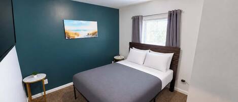 A hotel room with a bed, a small bedside table, a nightstand, a painting on the wall, and a window with curtains.