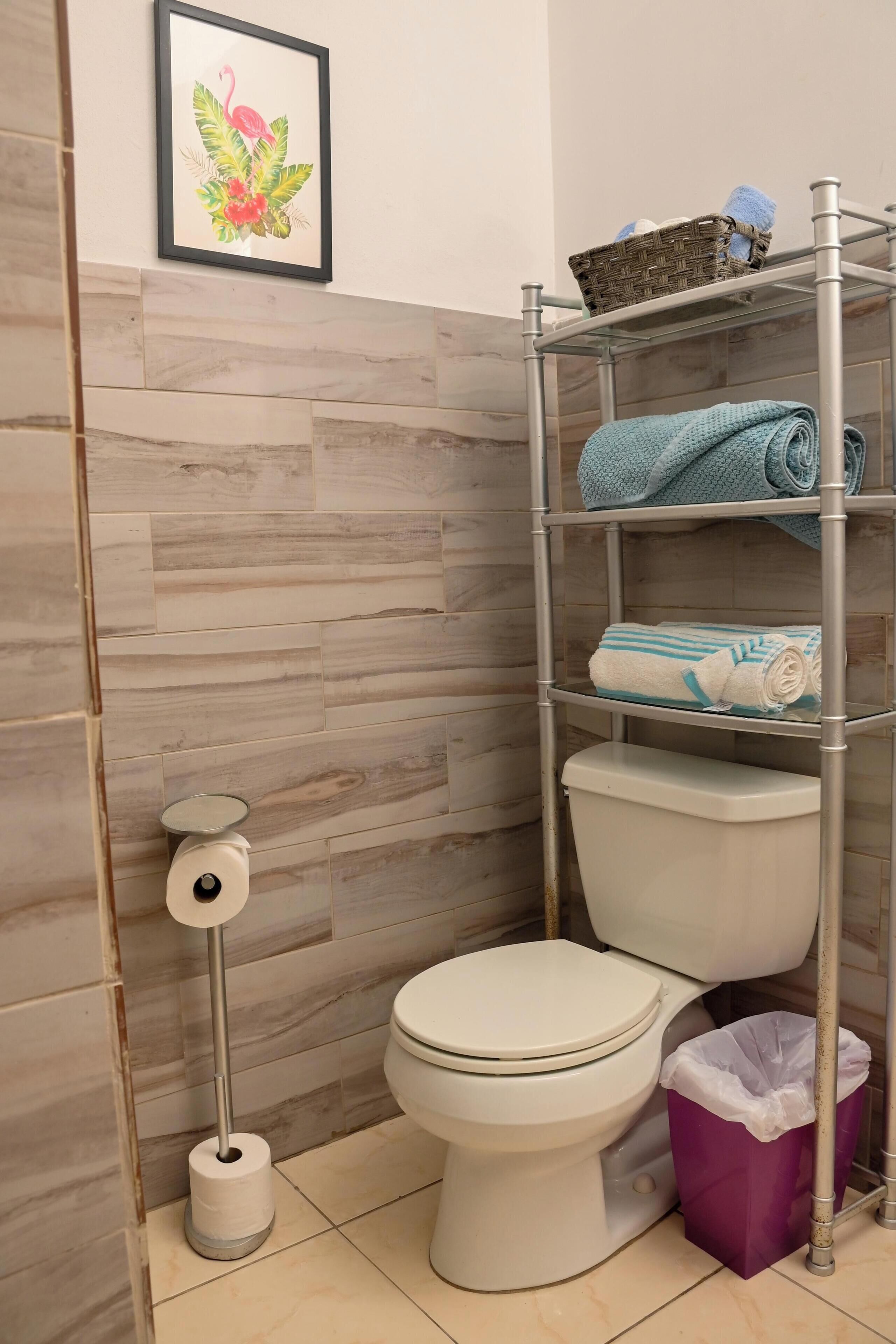 Un baño con un inodoro, un rollo de papel higiénico, un cubo de basura y un estante con toallas.