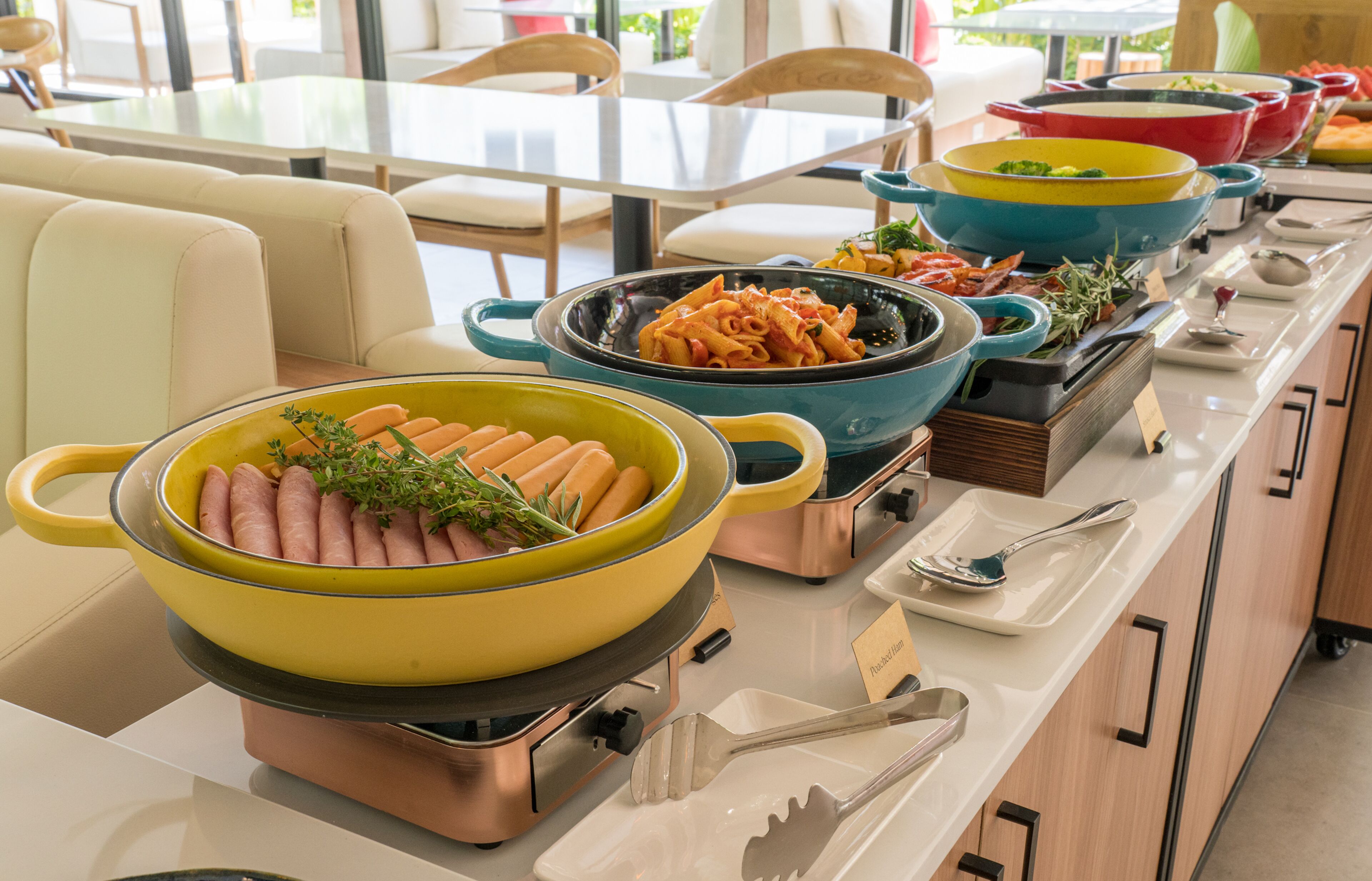 A buffet setup with various dishes, including a large yellow pot with sausages and a blue pot with pasta.