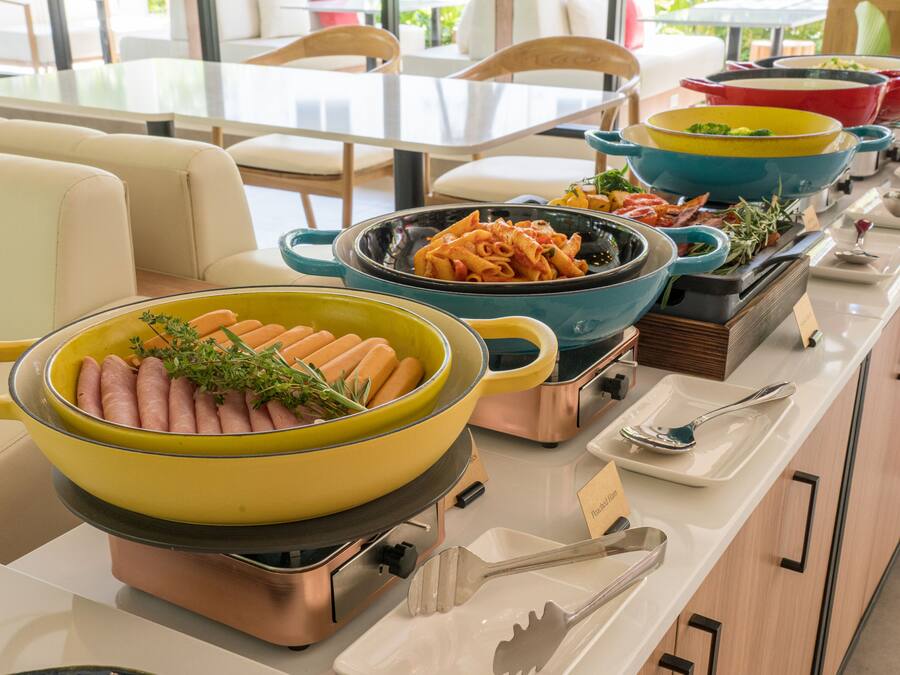 Montaje de bufé con diversos platos, incluyendo una olla amarilla grande con salchichas y una olla azul con pasta.