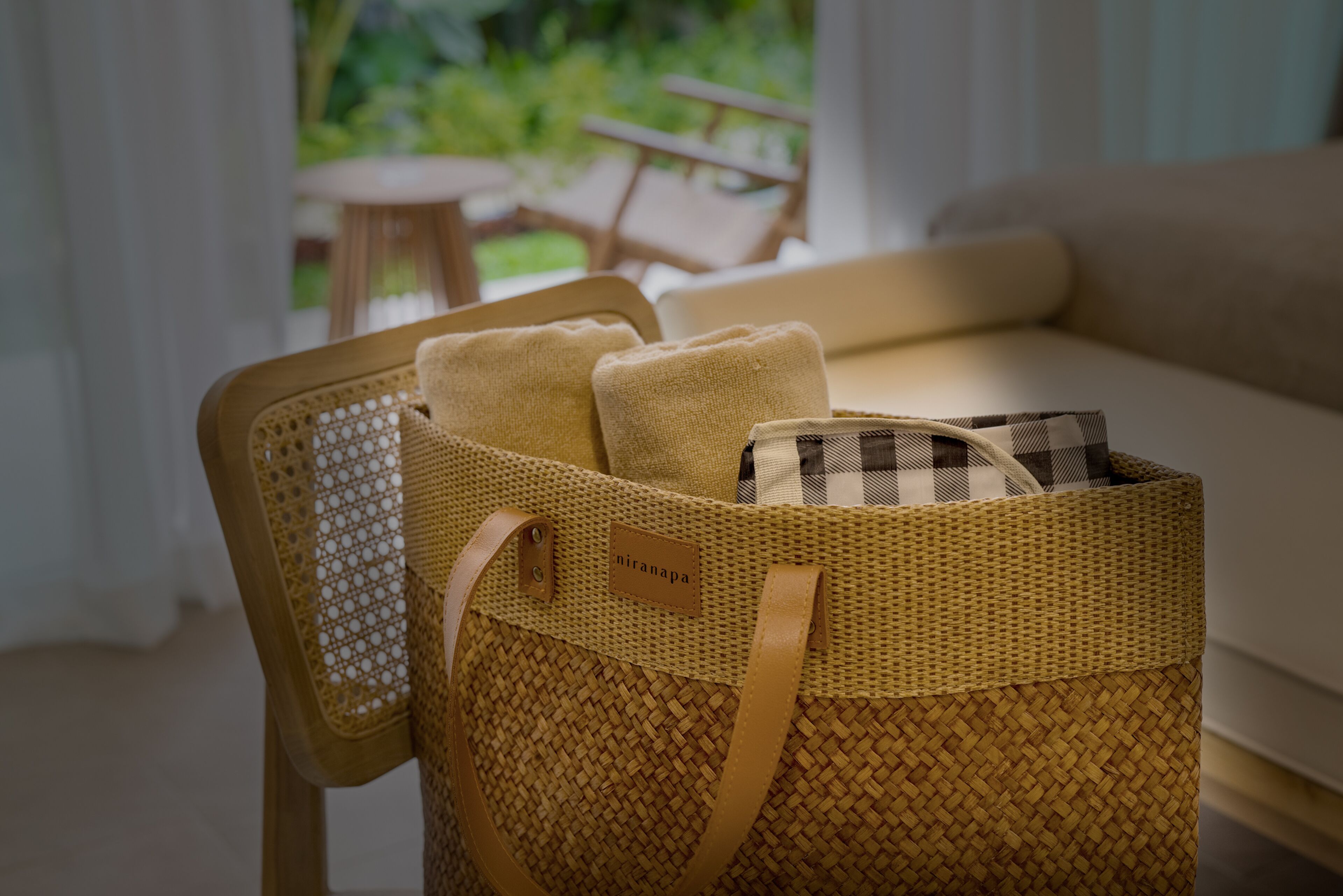 A wicker basket with a label, containing folded towels and a checkered item, placed on a chair near a sofa.