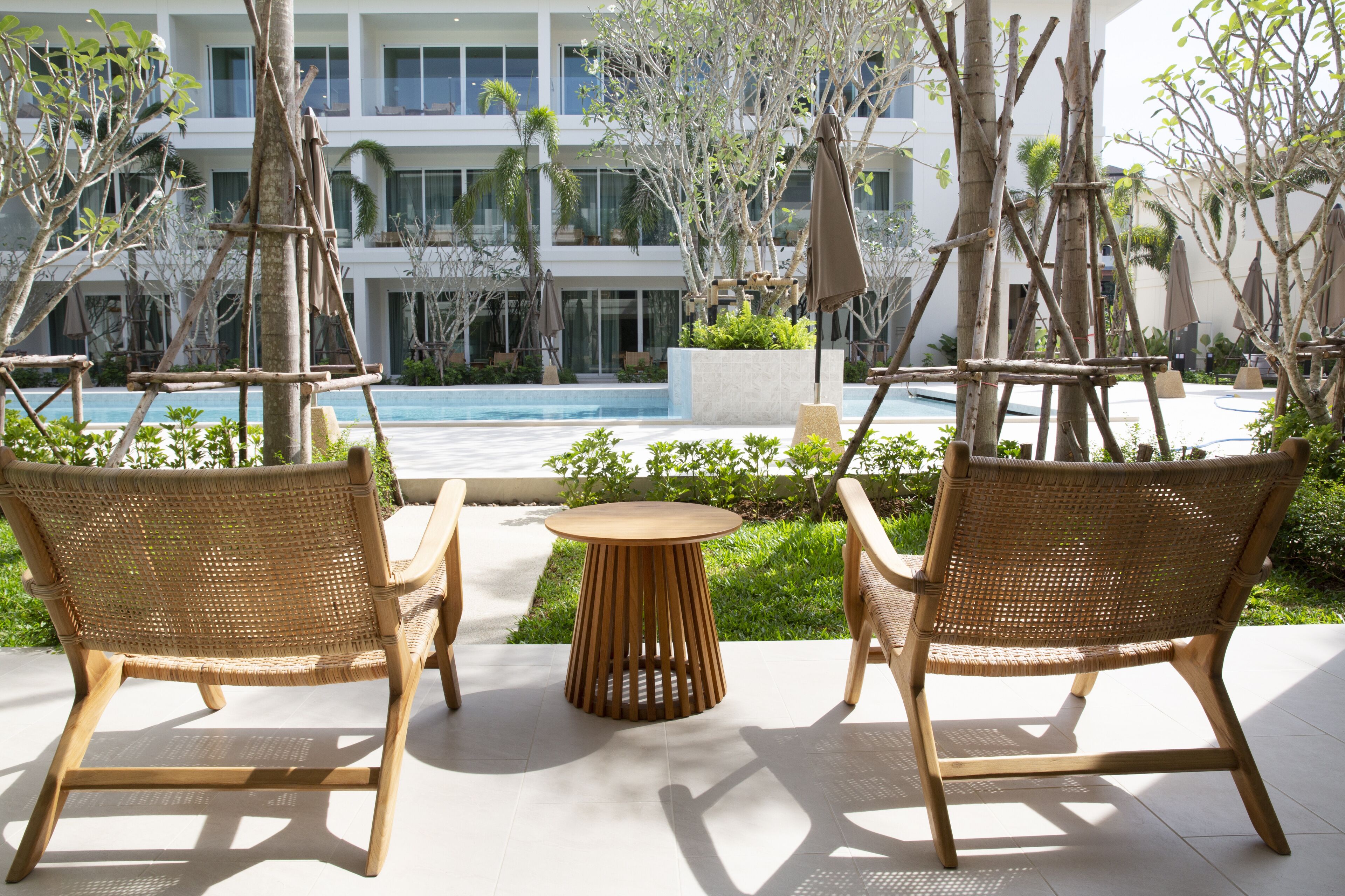 A poolside area with wicker chairs and a round table, surrounded by greenery and a building in the background.