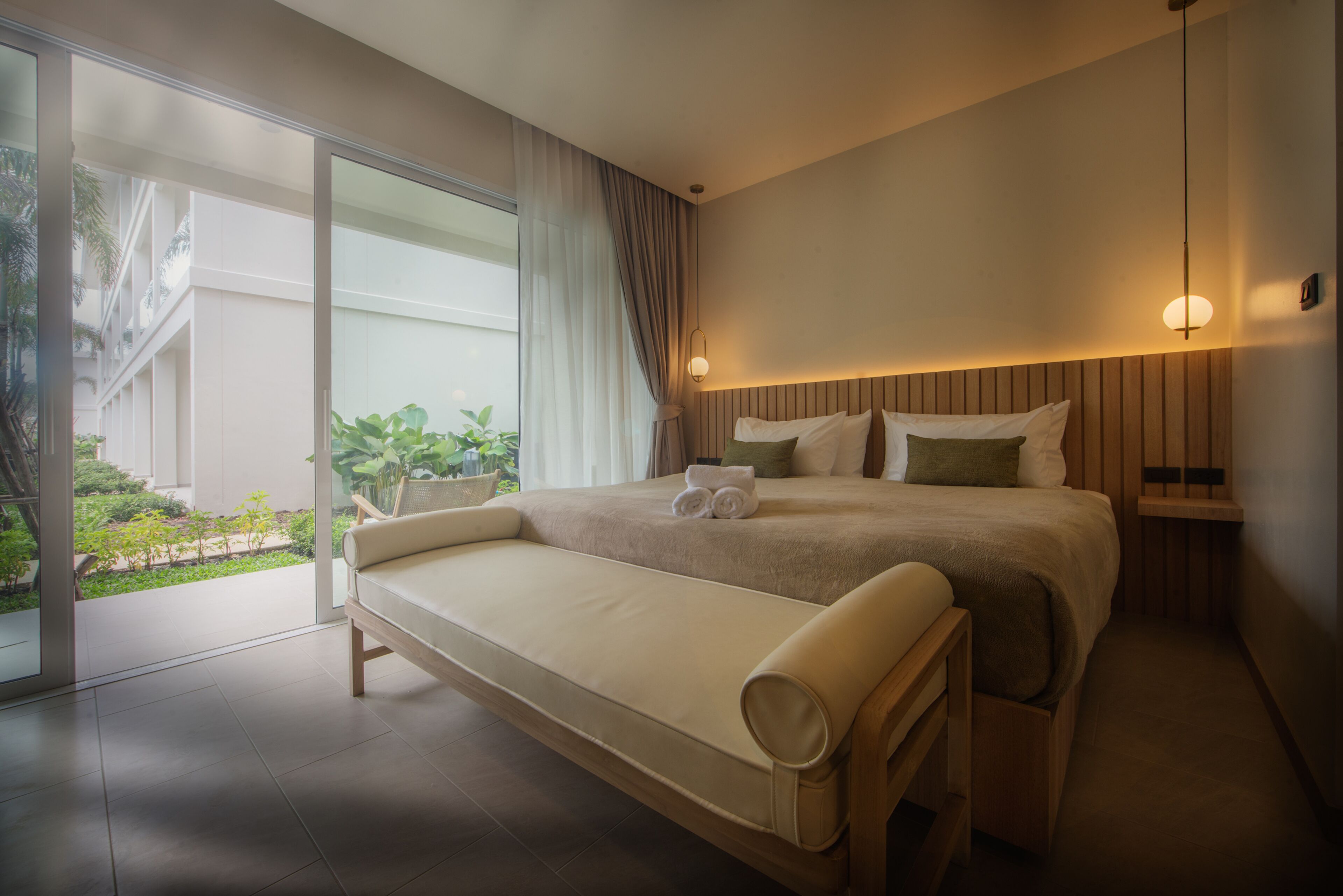 A modern hotel room with a large bed, a small sofa, and a sliding glass door leading to a balcony with plants.