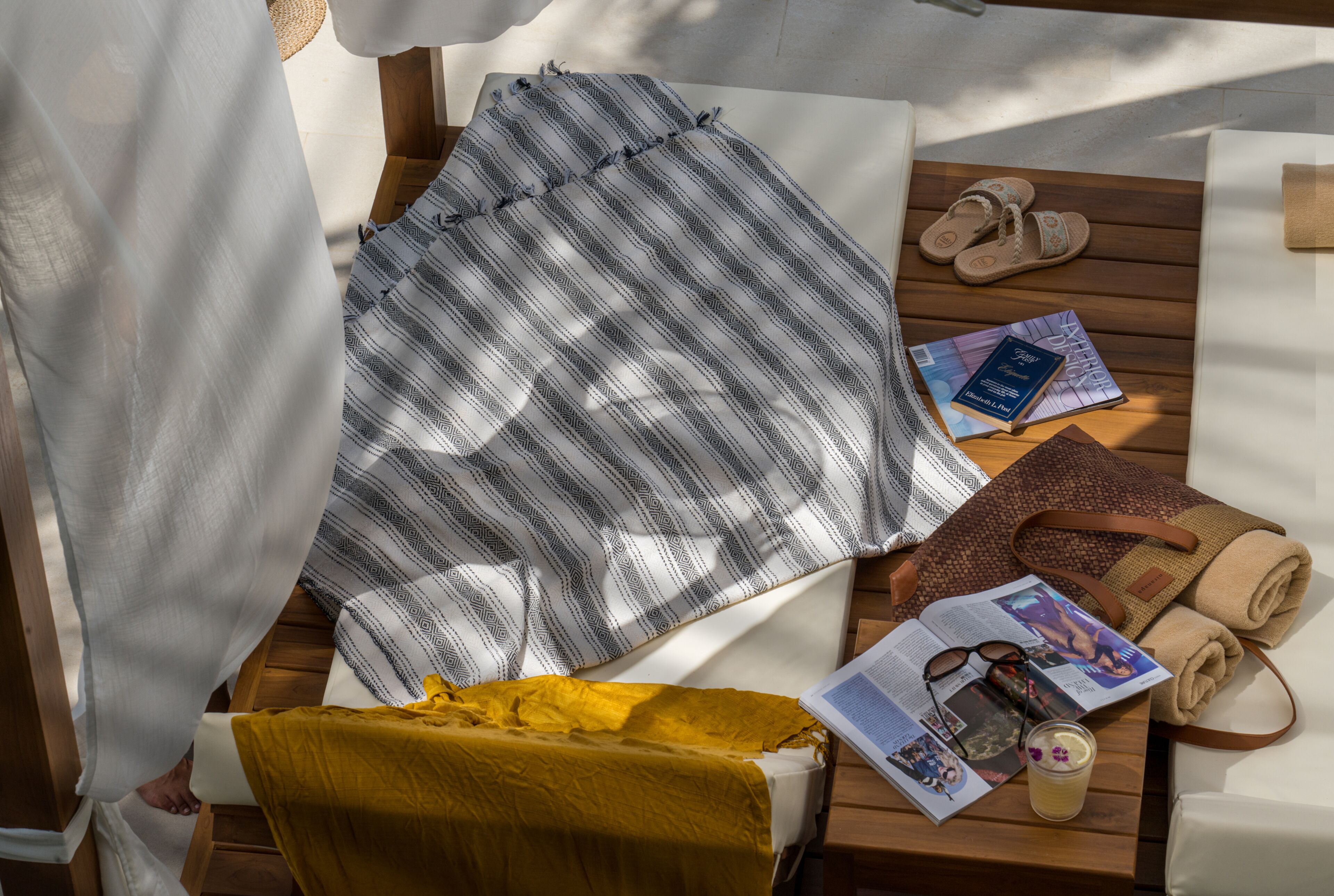 A sunlit room with a bed, a wooden table with a magazine, a pair of slippers, and a rolled-up towel.