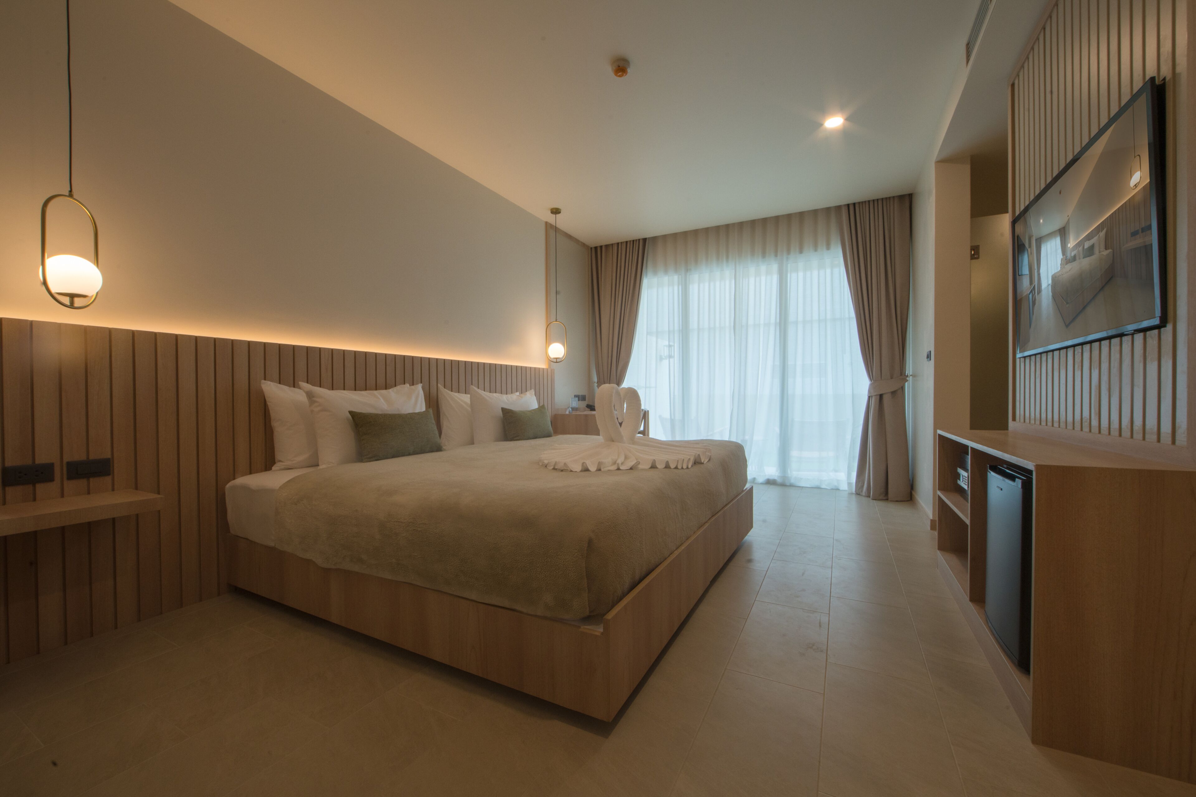 A modern hotel room with a large bed, wooden headboard, bedside tables, a built-in TV, and a large window with curtains.