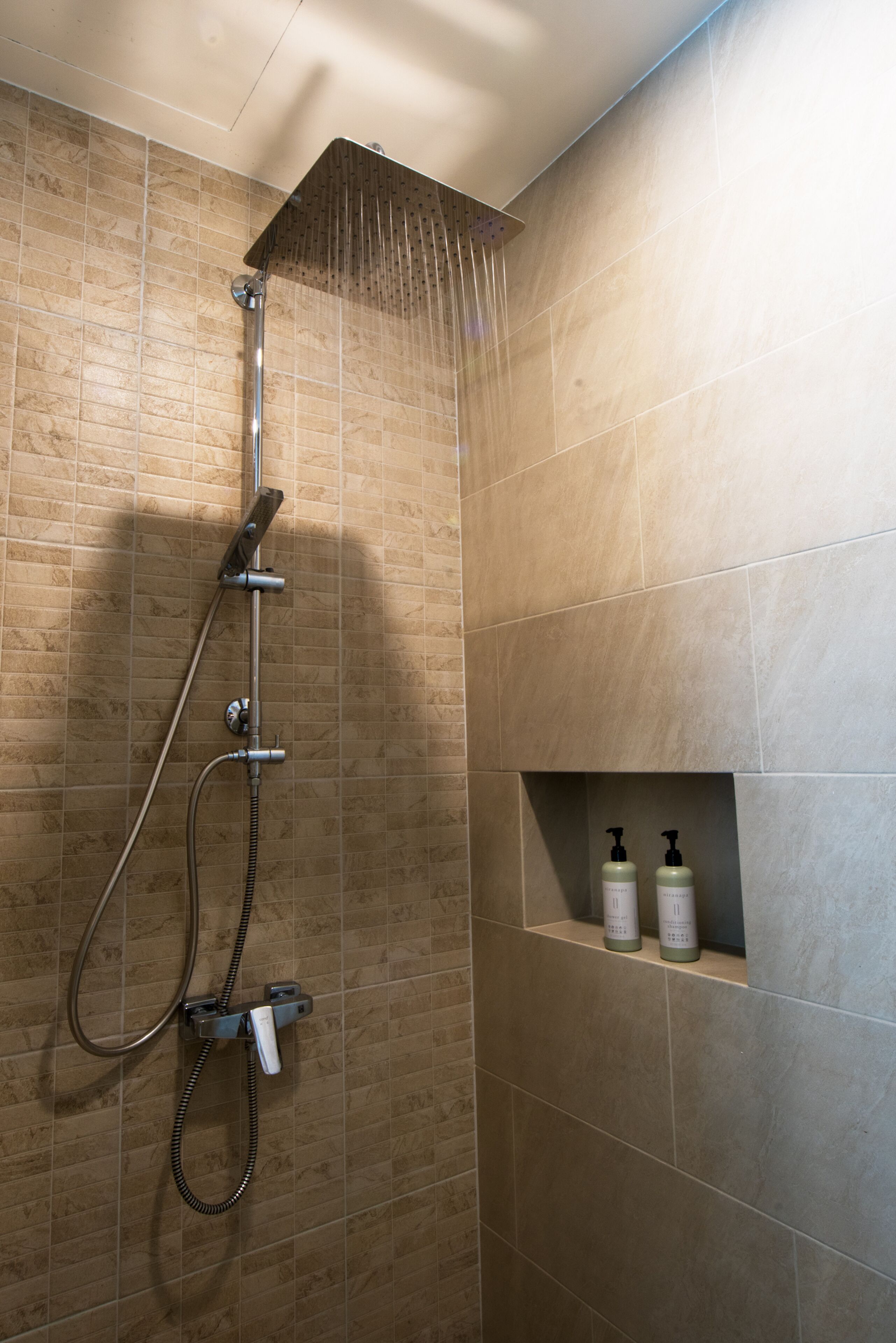 A shower with a handheld showerhead and a wall-mounted panel. There are two bottles of shampoo and conditioner on a shelf below the shower.