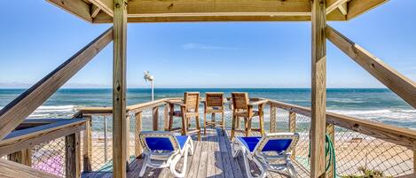 Una terraza de madera con dos sillas reclinables y una mesa, con vista a una playa y una vista despejada del océano.