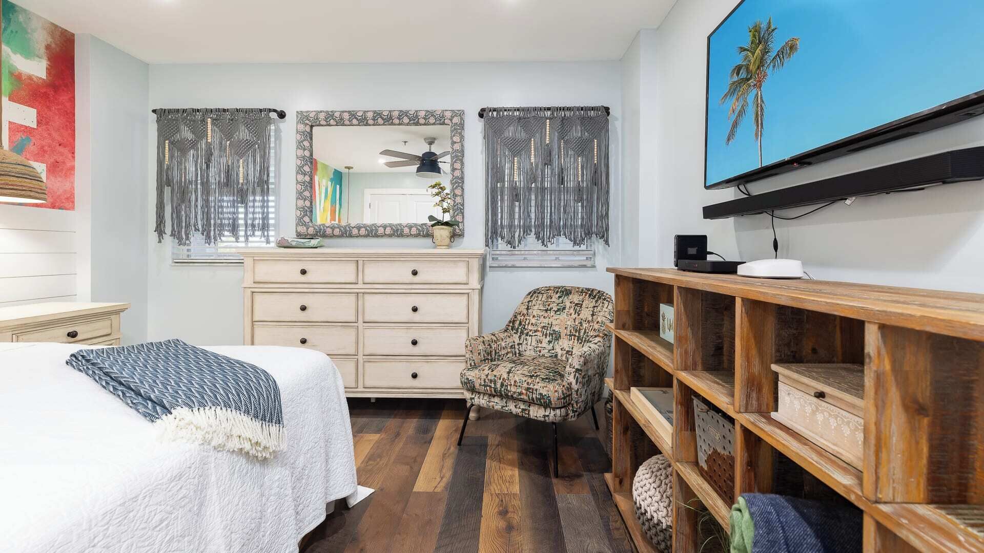 Quarto com cama, cadeira, penteadeira, espelho e televisão sobre uma estante de madeira.