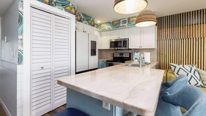 Una cocina con armarios blancos, un microondas y una estufa. Hay un banco azul con un cojín y una almohada estampada.