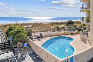 A swimming pool with a view of the beach, surrounded by lounge chairs and a fenced deck area.