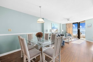 A dining area with a glass table and chairs, a ceiling fan, and a view of the beach through an open door.