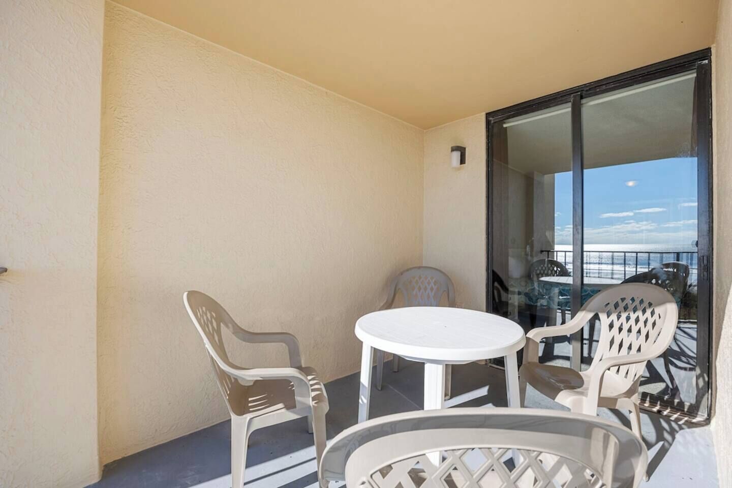 Un balcón con una mesa blanca y cuatro sillas de plástico, una puerta de vidrio que da a un balcón con vistas, y un pequeño aplique de pared.