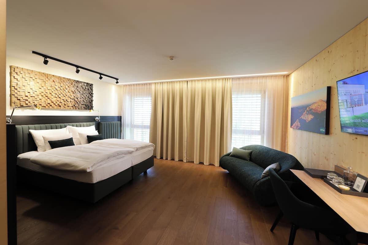 A hotel room with a bed, a sofa, a desk with a chair, and a TV on the wall.