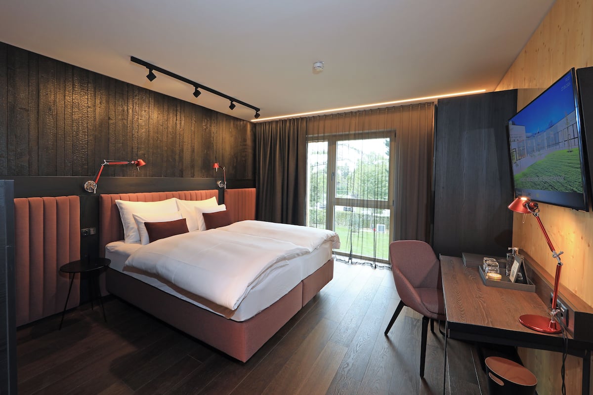 A modern hotel room with a large bed, a desk with a chair, a television, and a view of the outdoors.