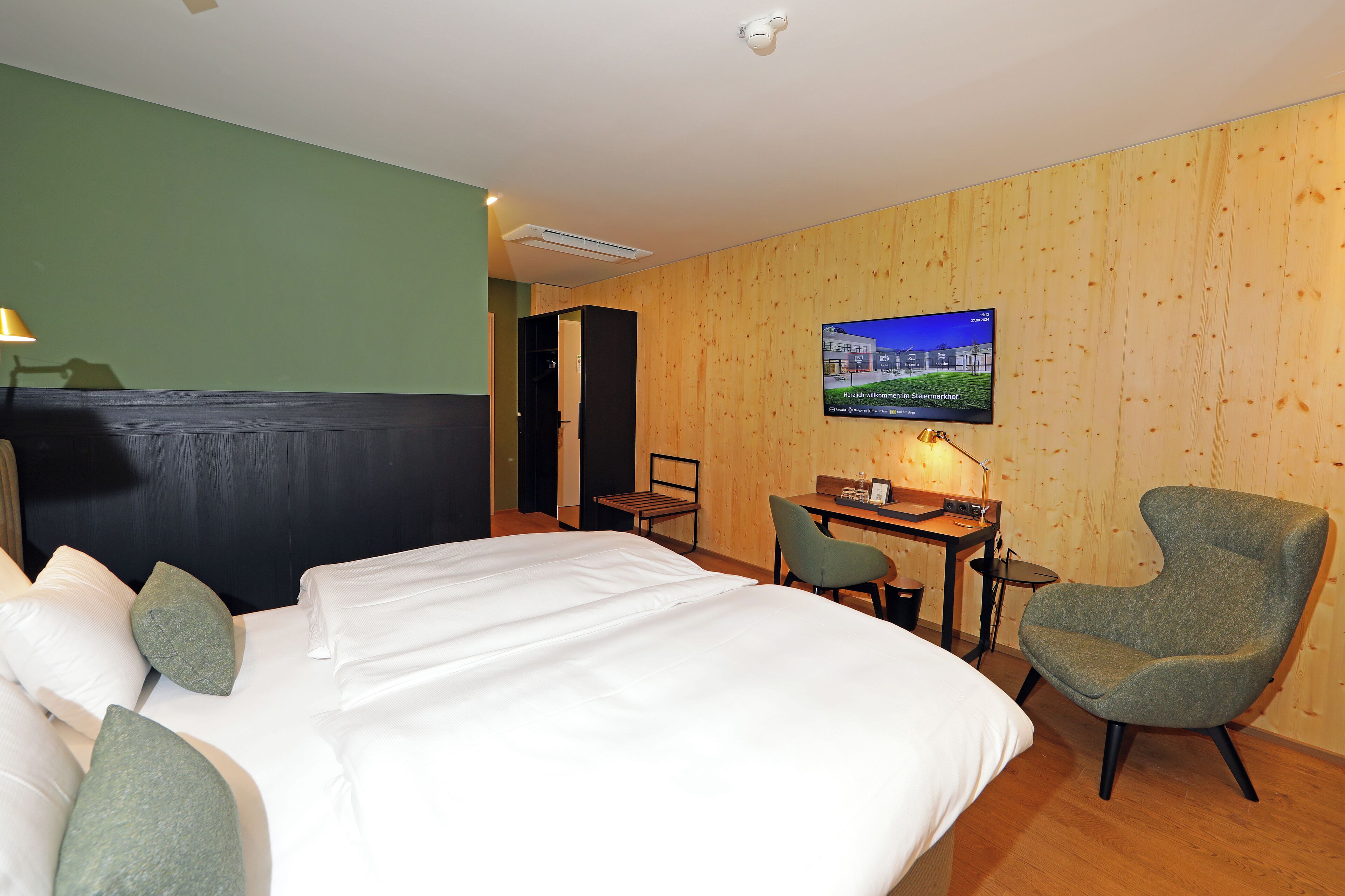 A hotel room with a bed, two chairs, a desk, a television, and a wooden wall.