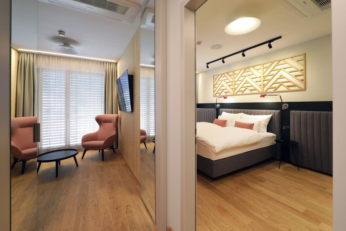 A modern hotel room with a large bed, a TV, a desk, and a seating area with chairs.