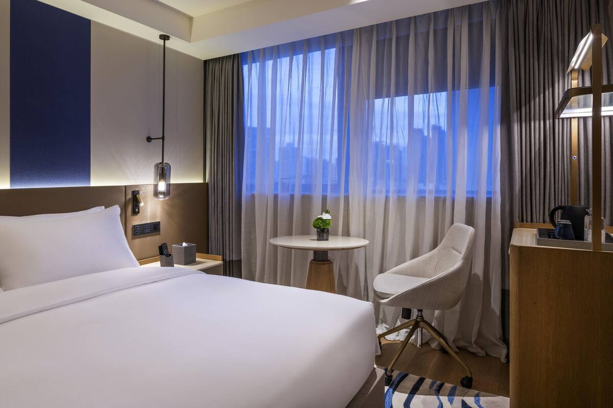 A hotel room with a large bed, a desk with a chair, a small table with a plant, and a view of the city through sheer curtains.