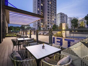 An outdoor seating area with tables and chairs, a covered section with blue lighting, and a view of a cityscape with buildings in the background. - Novotel Shanghai Jingan (Shanghai)