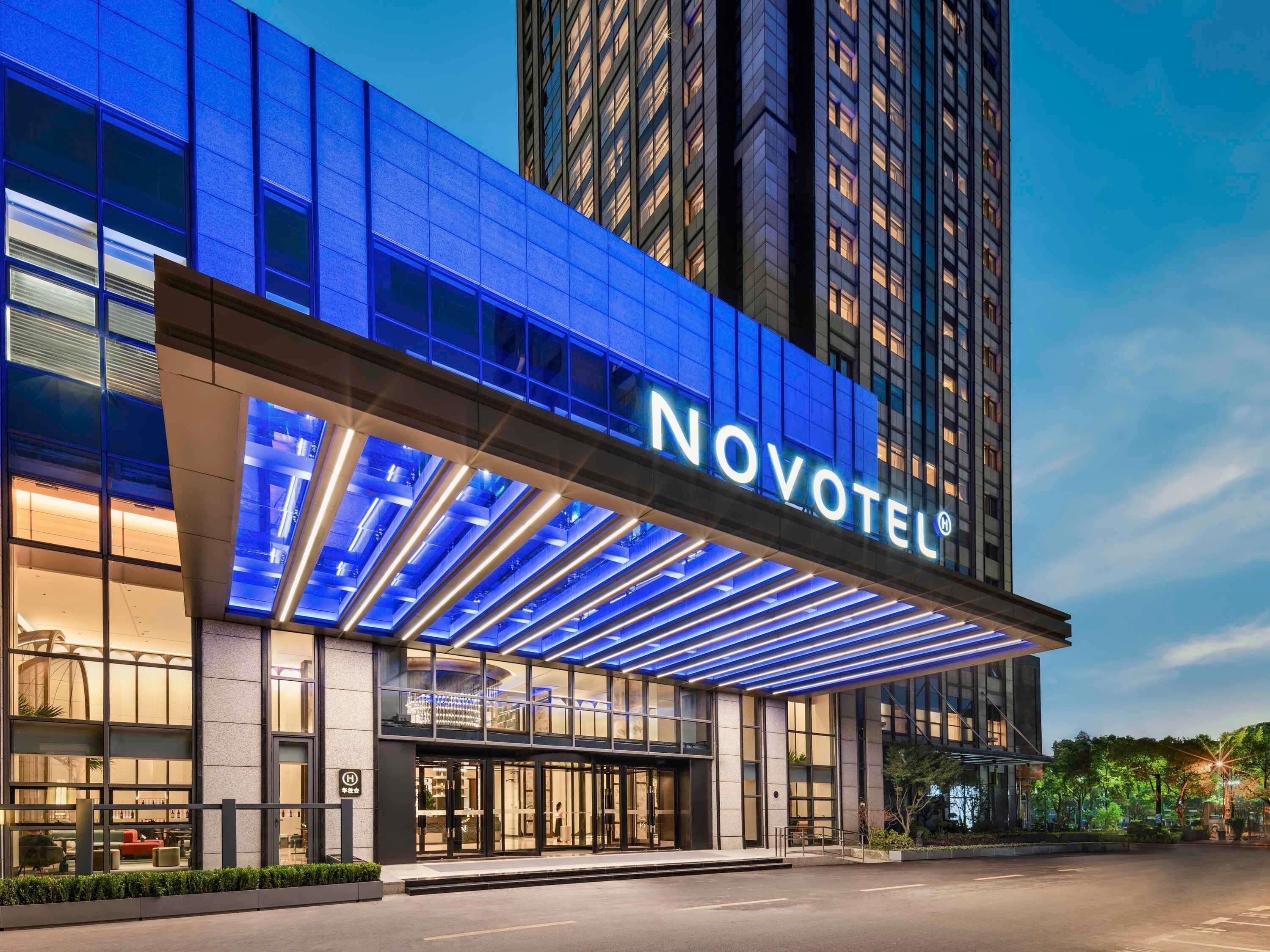 A hotel entrance with a prominent 'NOVOTEL' sign, a modern glass facade, and a well-lit entrance area.
