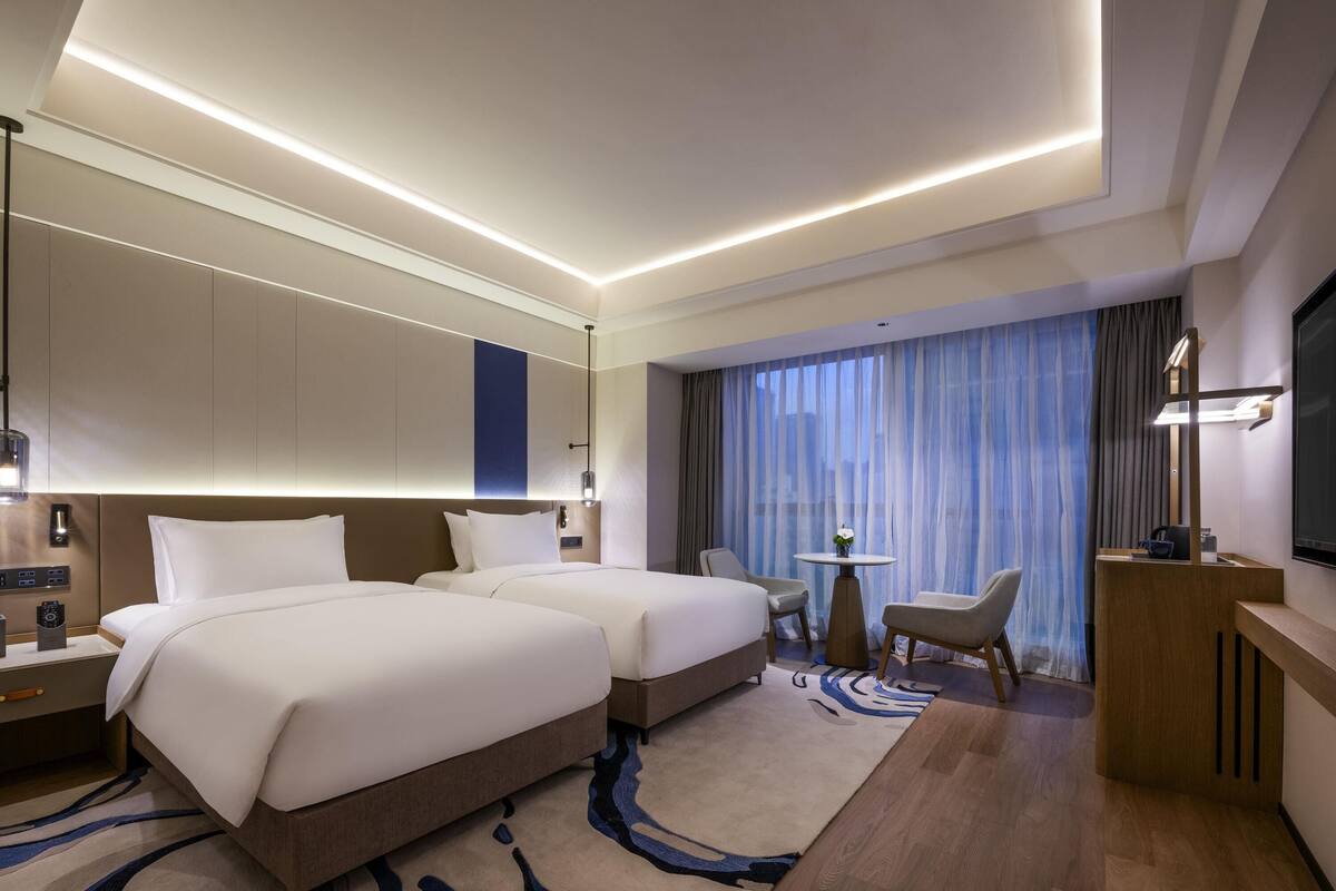 A hotel room with two beds, a desk, a chair, a TV, and a large window with curtains.